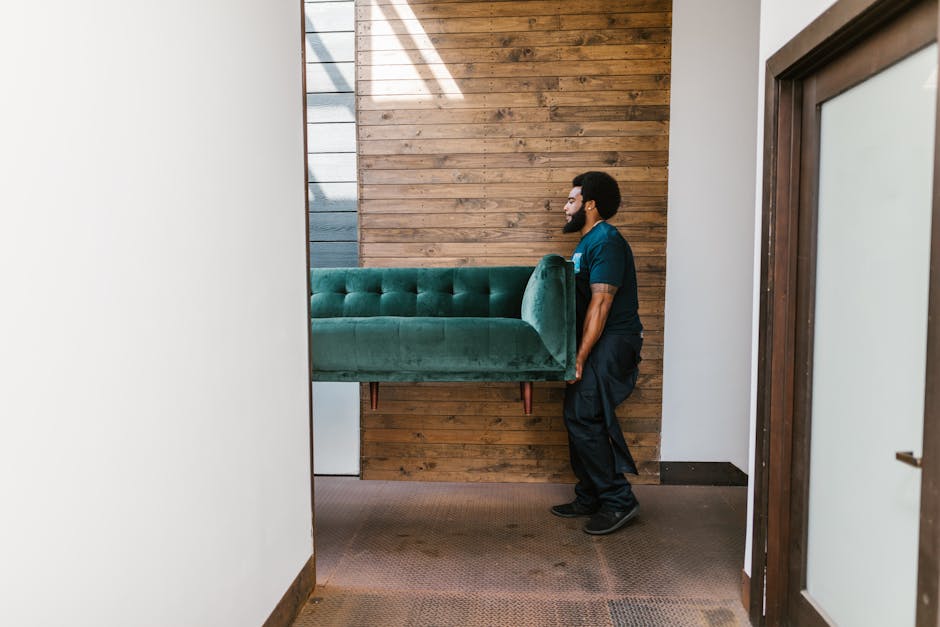 A professional mover from France Removals is carefully lifting a green upholstered sofa with tufted buttons and wooden legs through an interior doorway during a home relocation process. The mover is w