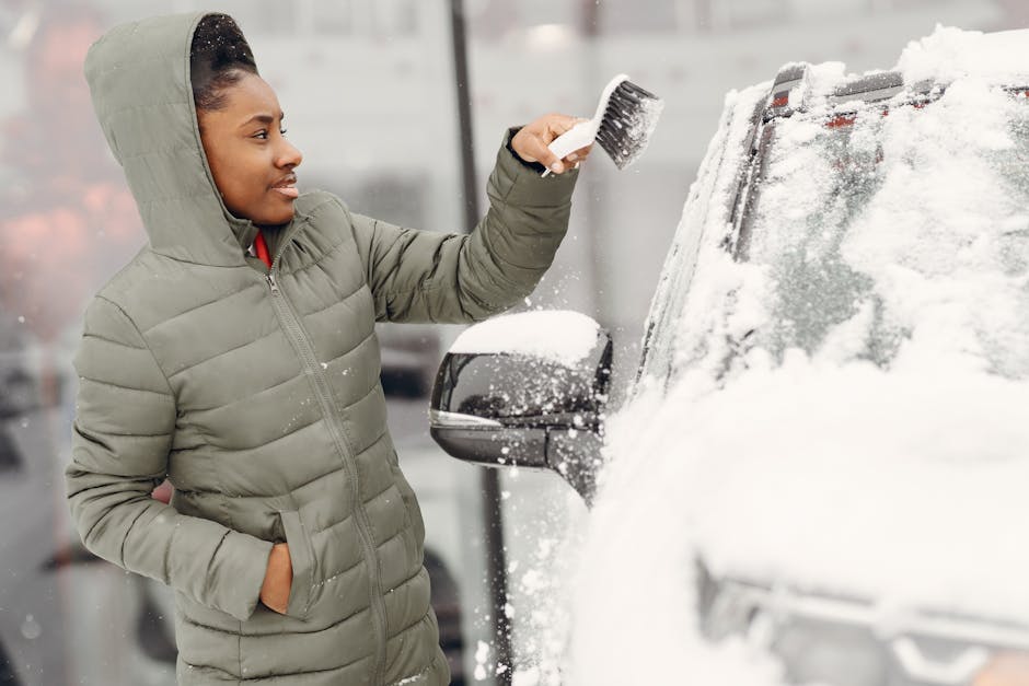 A woman wearing a green padded winter jacket with a hood, standing outdoors next to a vehicle covered in snow, is using a small brush to clear snow from the vehicle's side mirror. The vehicle appears 